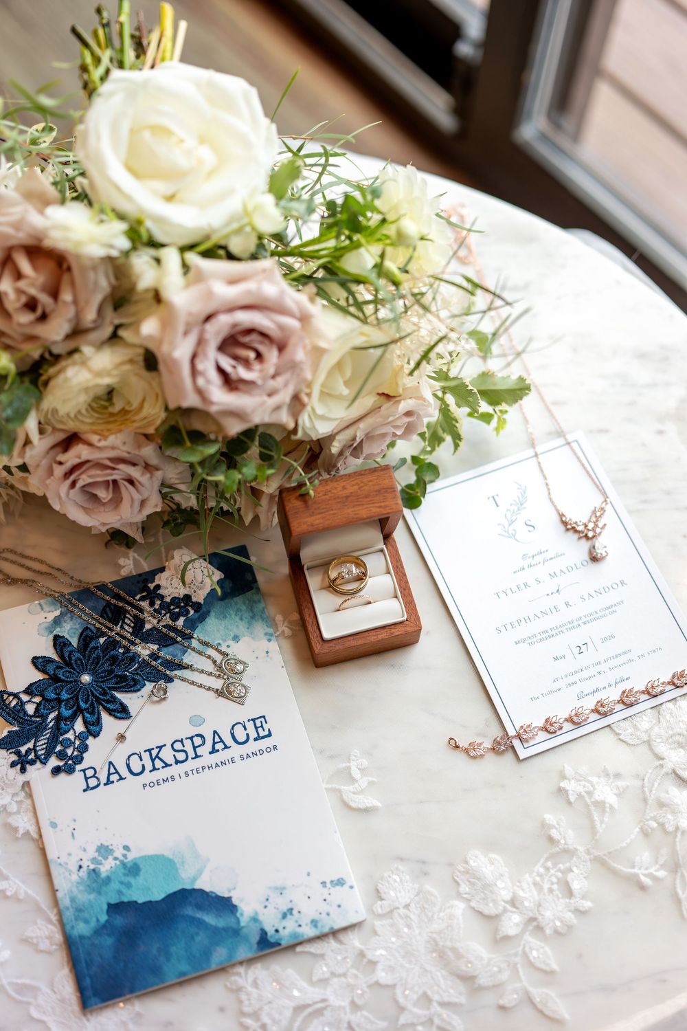 Elegant wedding flat lay with blush roses bouquet, gold rings in wooden box, invitation, jewelry, and Backspace poetry book on white lace.