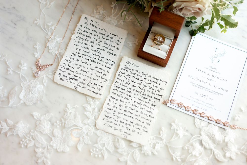 Wedding flat lay with handwritten vows, gold rings in wooden box, lace fabric, rose gold bracelet, and elegant invitation.