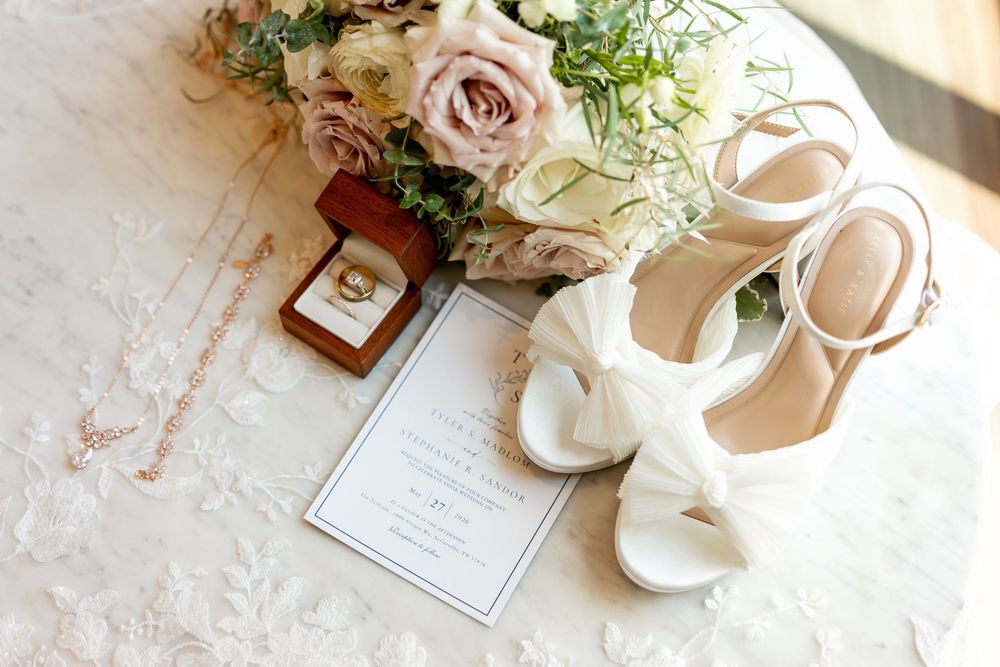 Elegant wedding flat lay with white heels, floral bouquet, gold ring box, invitation, and delicate necklace on lace fabric.