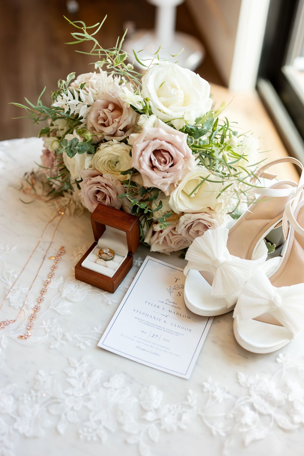 Elegant bridal flat lay with blush roses bouquet, white heels, wedding rings, invitation, and delicate gold jewelry on lace.