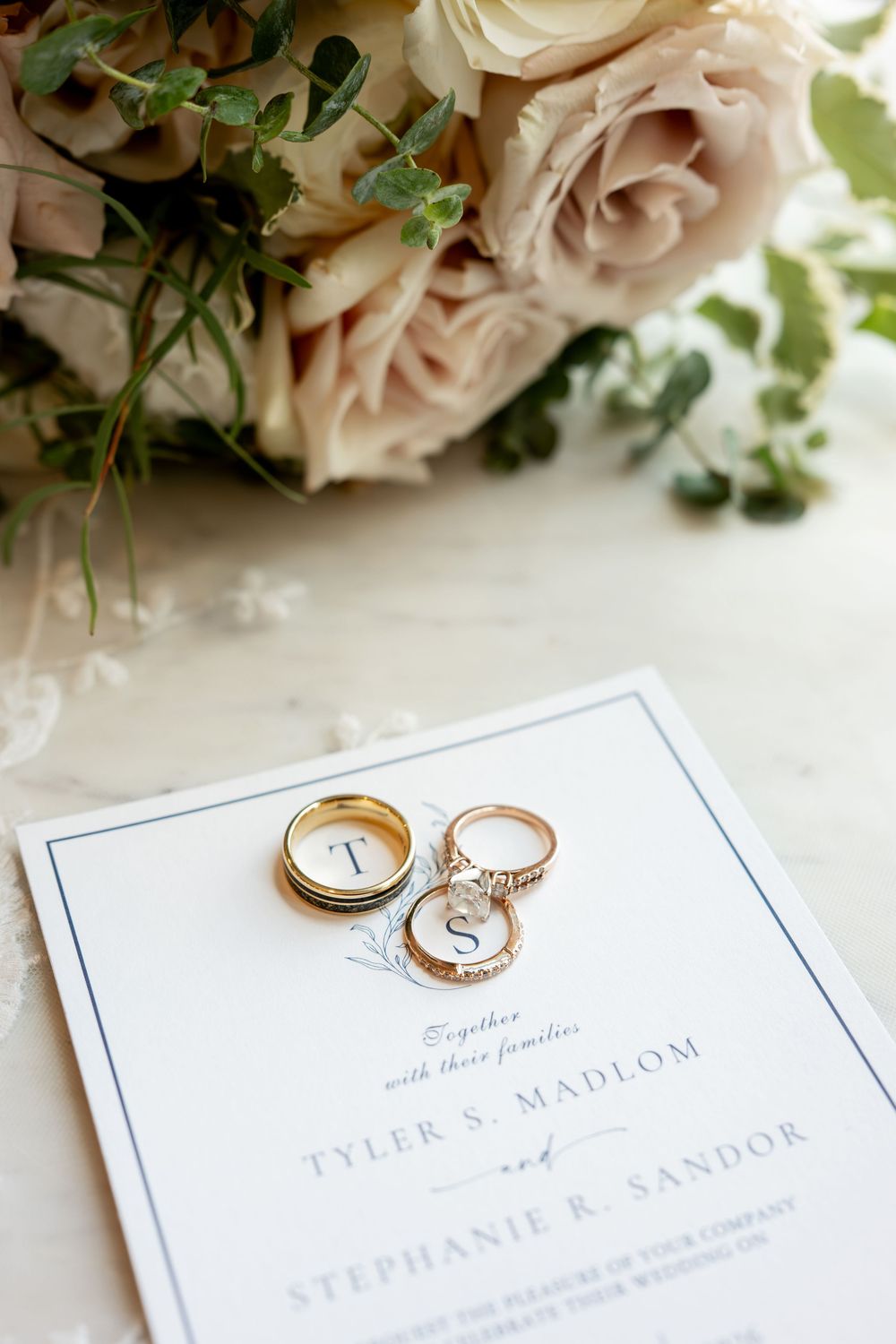 Wedding rings and engagement ring rest on an elegant invitation for Tyler S. Madlom and Stephanie R. Sandor beside blush roses.