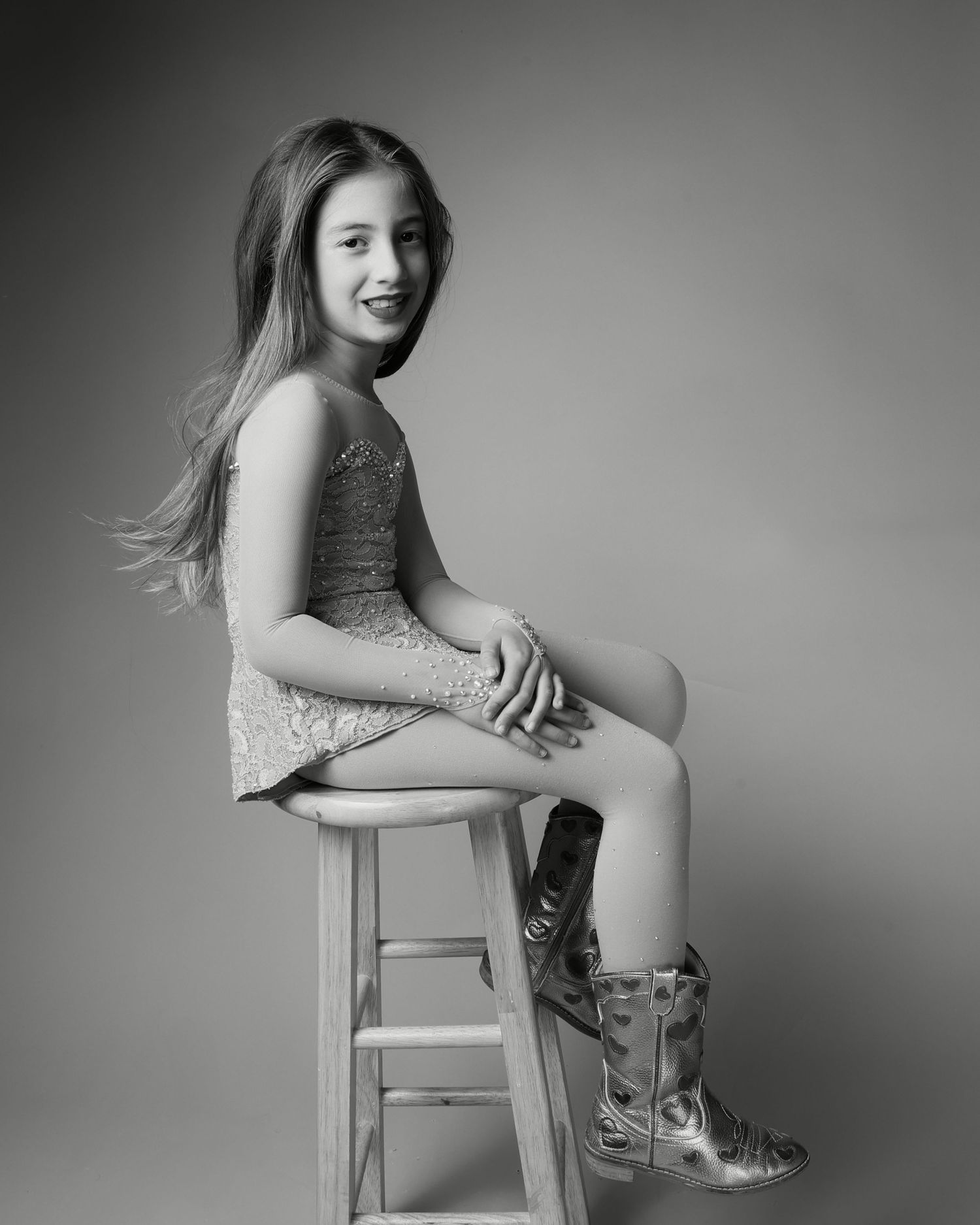 Black and white studio portrait of a young girl sitting on a stool, wearing a sparkly dress and cowboy boots, smiling.