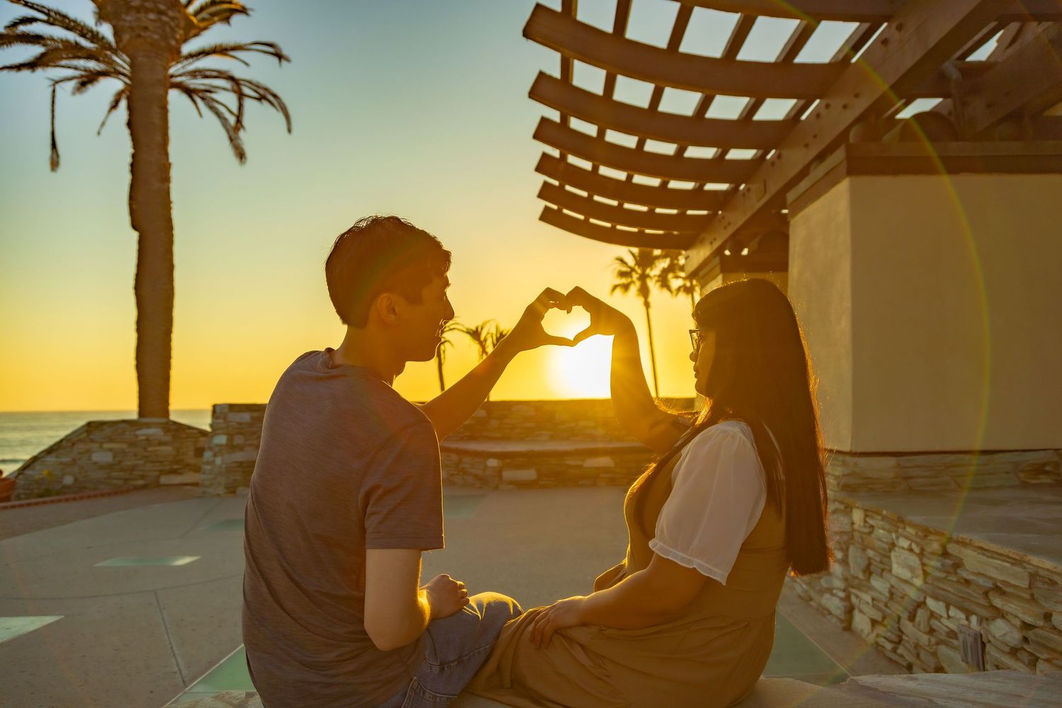 Couple making heart shape with hands at sunset near palm trees and ocean.