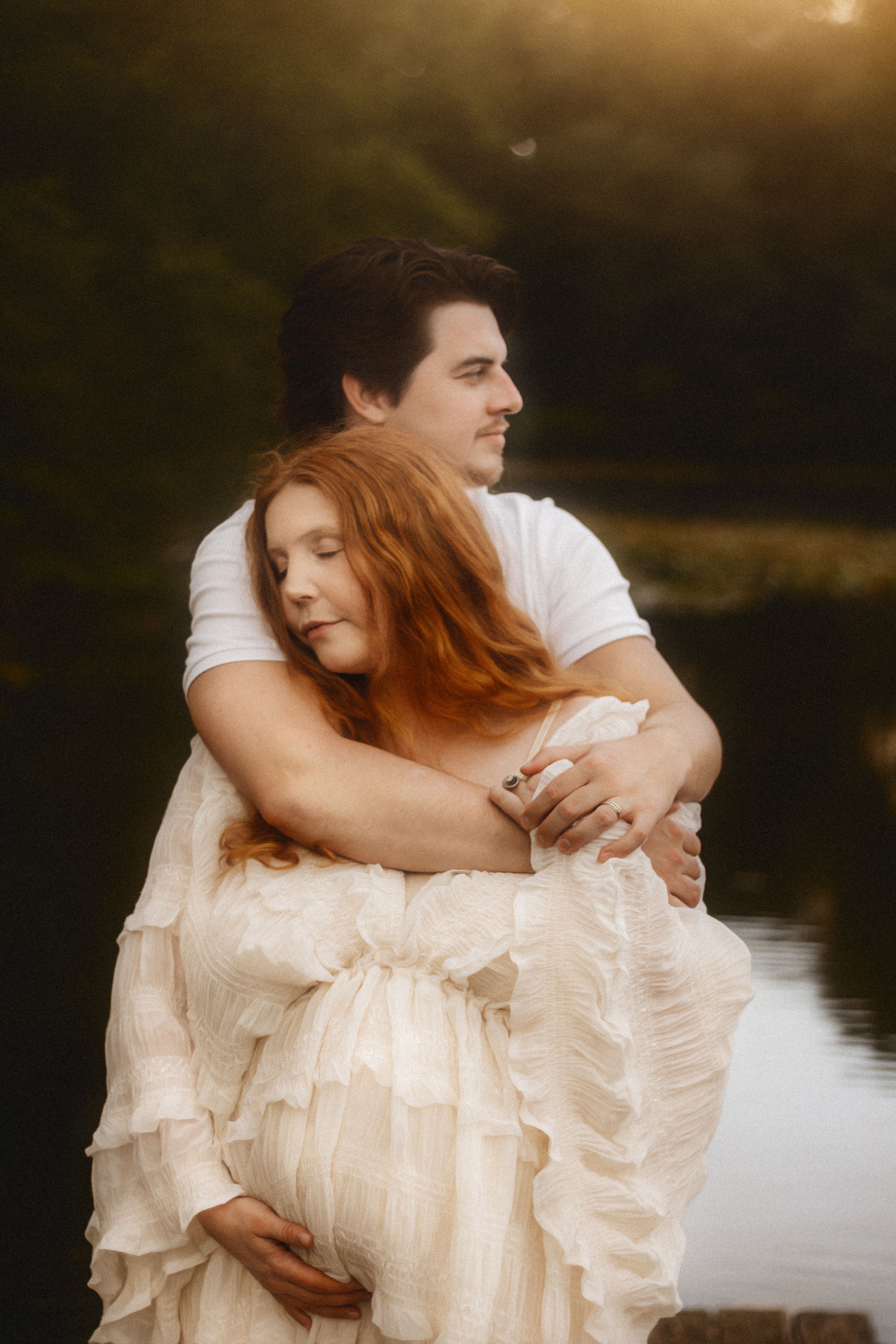 A couple shares a tender embrace outdoors at golden hour, the woman in a flowing white gown cradling her baby bump.