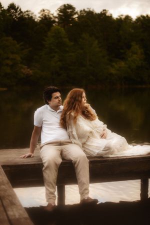 Couple in white sitting on a wooden dock by a lake, surrounded by lush green trees.