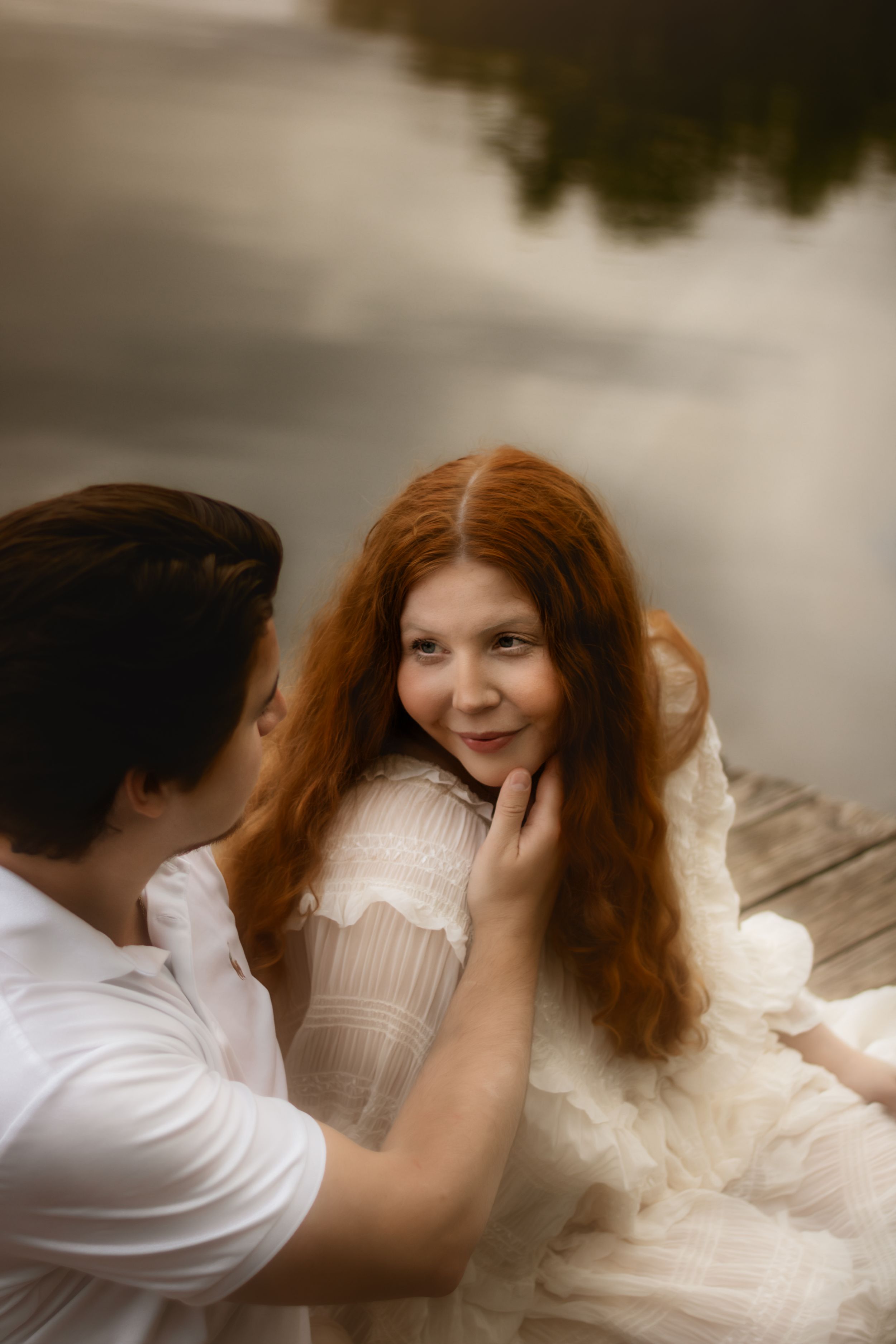 A man tenderly touches a redhead woman's face as they sit together on a dock by a misty lake.