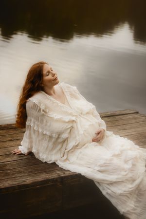 Pregnant woman in white ruffled dress sitting on wooden dock by calm lake with long red hair and eyes closed.