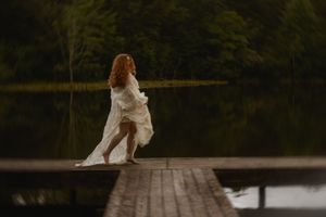 A red-haired woman in a flowing white dress stands on a wooden dock surrounded by a serene forest lake.