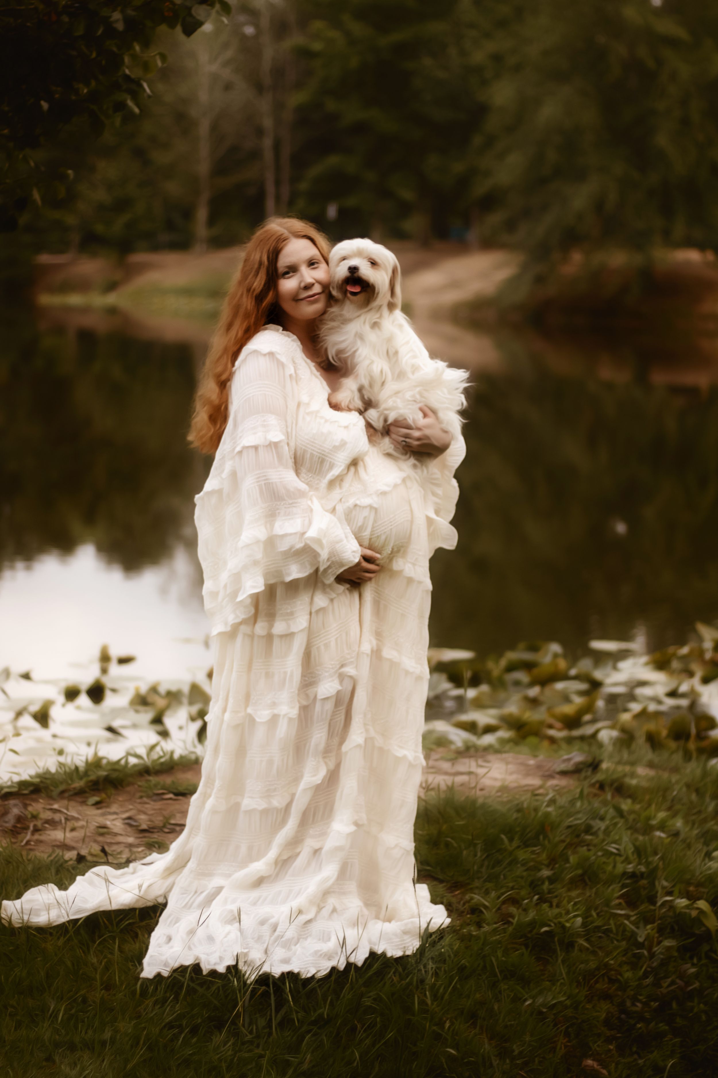 Pregnant woman in white ruffled dress holding small white dog by a serene pond surrounded by lush greenery.