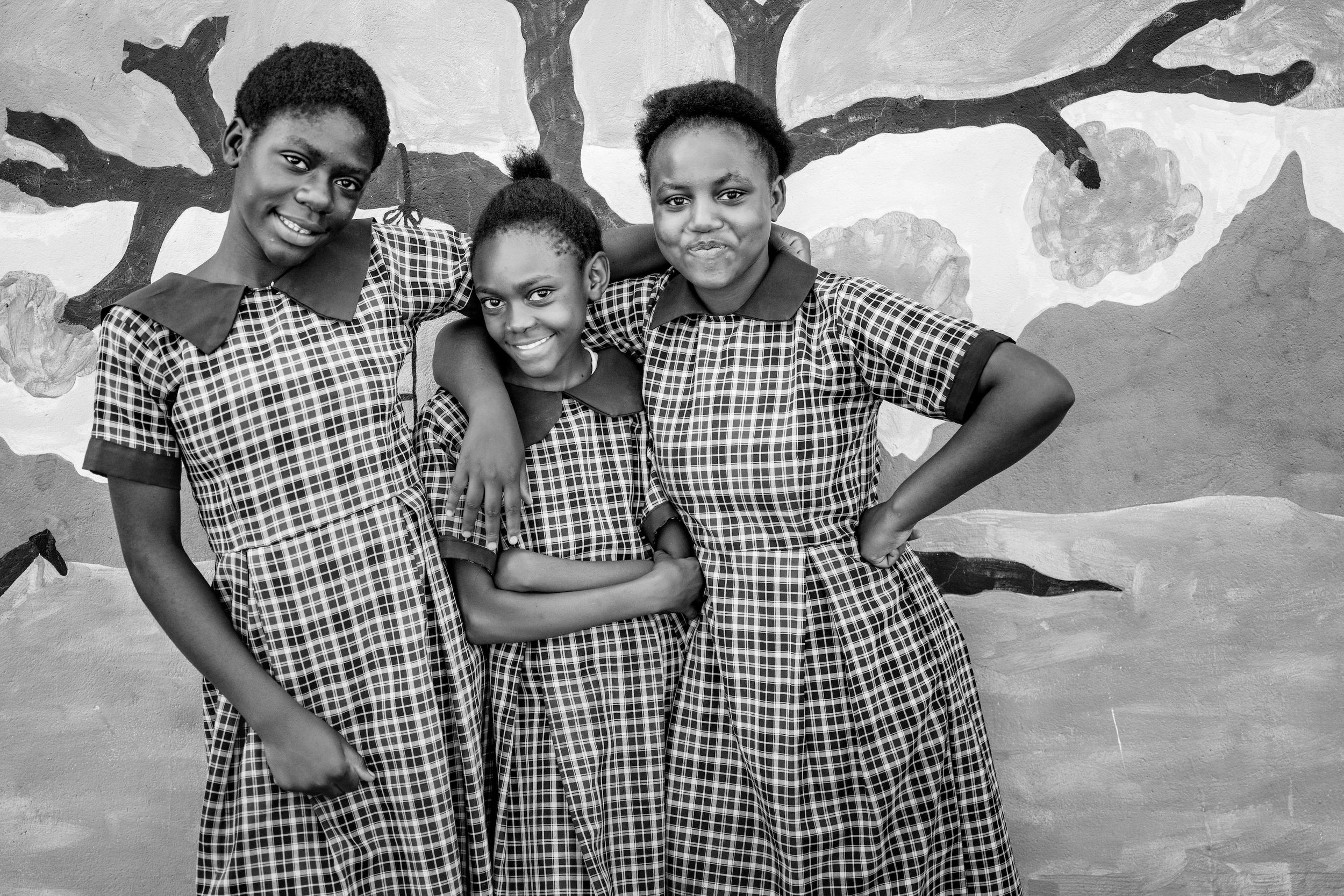 Three girls in matching plaid dresses pose together smiling in a black and white photo before a painted mural.