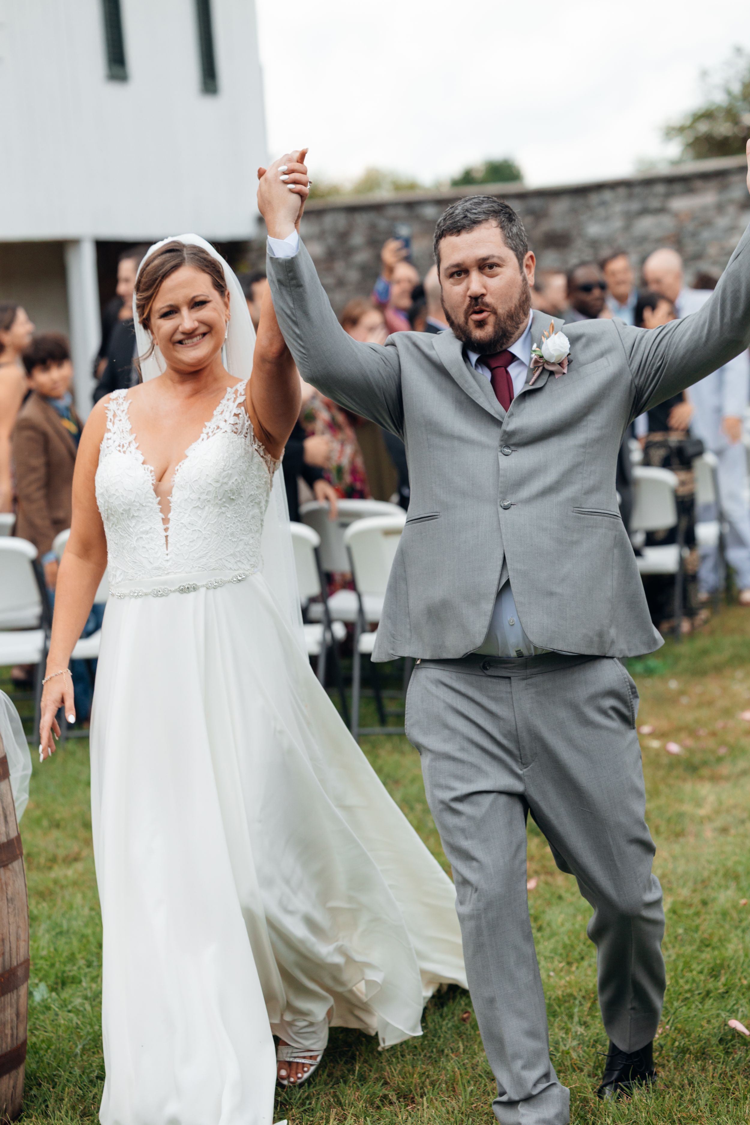 Joyful newlyweds celebrate after outdoor wedding ceremony, bride in lace gown and groom in gray suit raising hands.