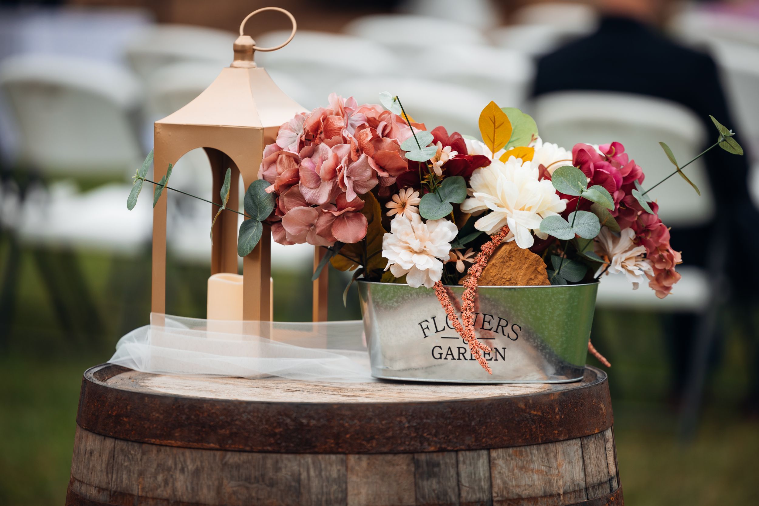 Rustic wedding decor with a lantern, pink hydrangeas, and colorful blooms in a tin on a wooden barrel.