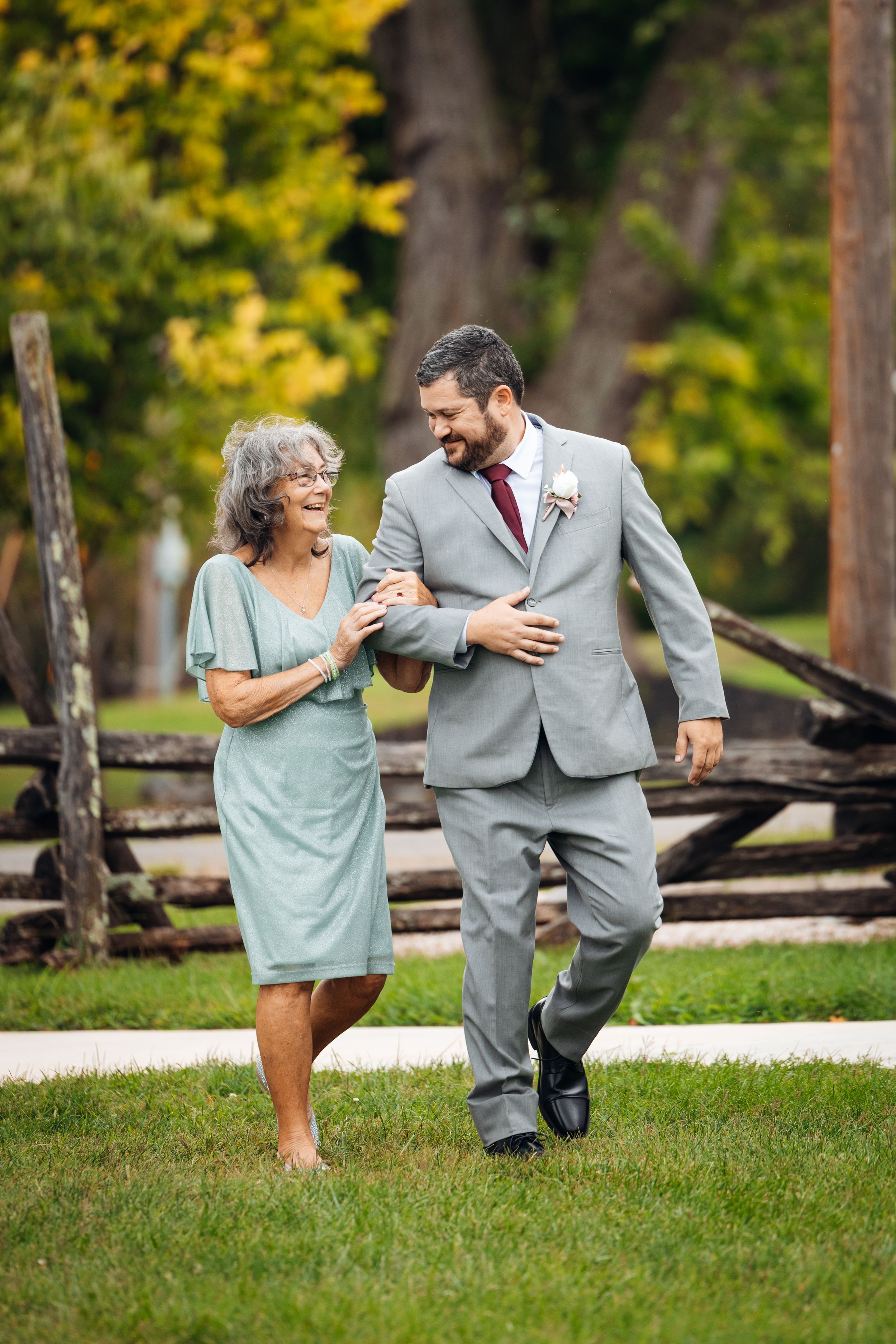 Groom in gray suit walks arm-in-arm with smiling older woman in sage dress at outdoor wedding venue.