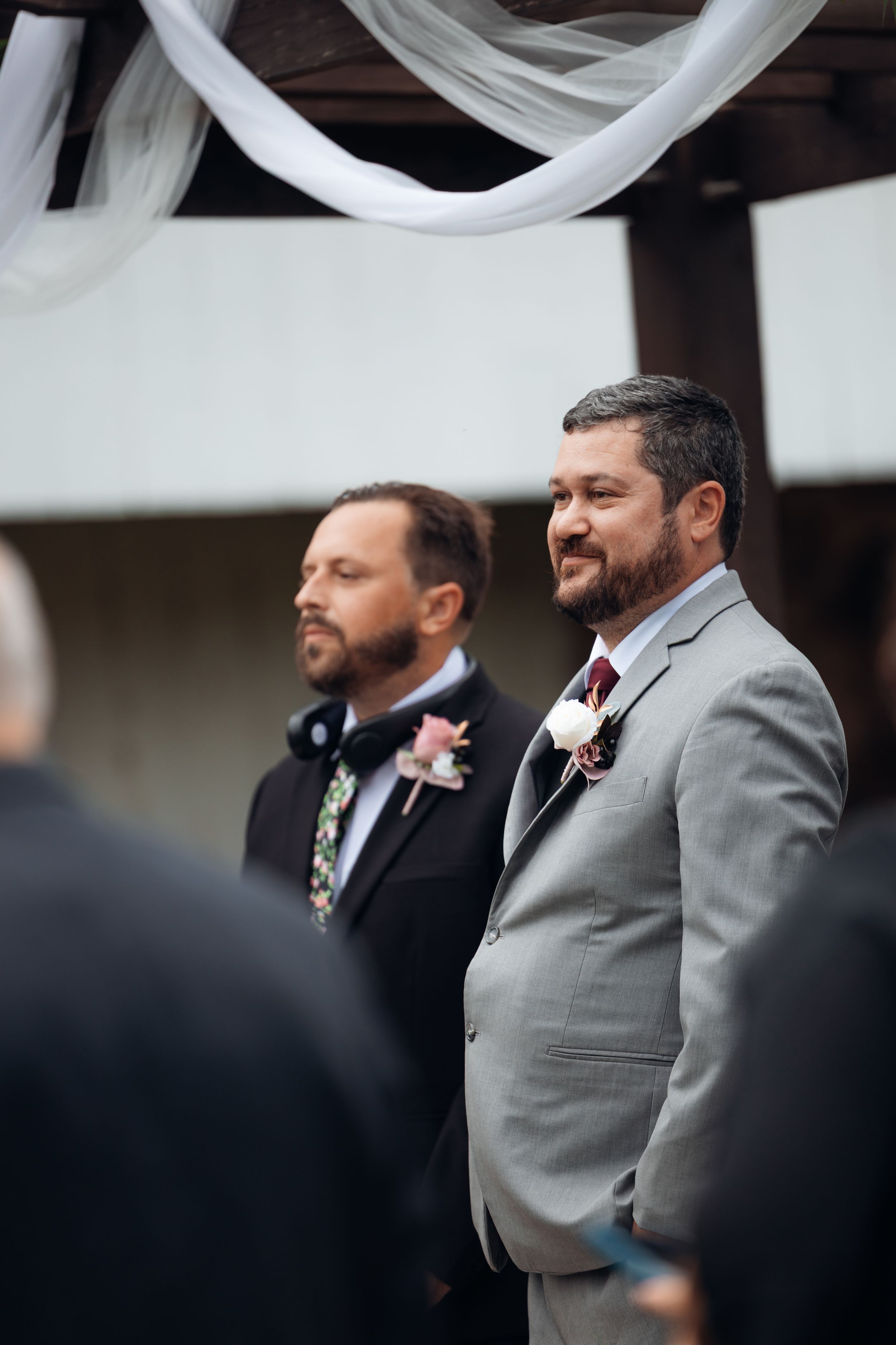 Groom in gray suit with boutonniere stands at wedding altar, emotional, with groomsman in black suit behind him.