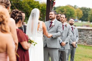 Outdoor wedding ceremony with groom in gray suit, groomsmen, and bridesmaids in burgundy dresses.
