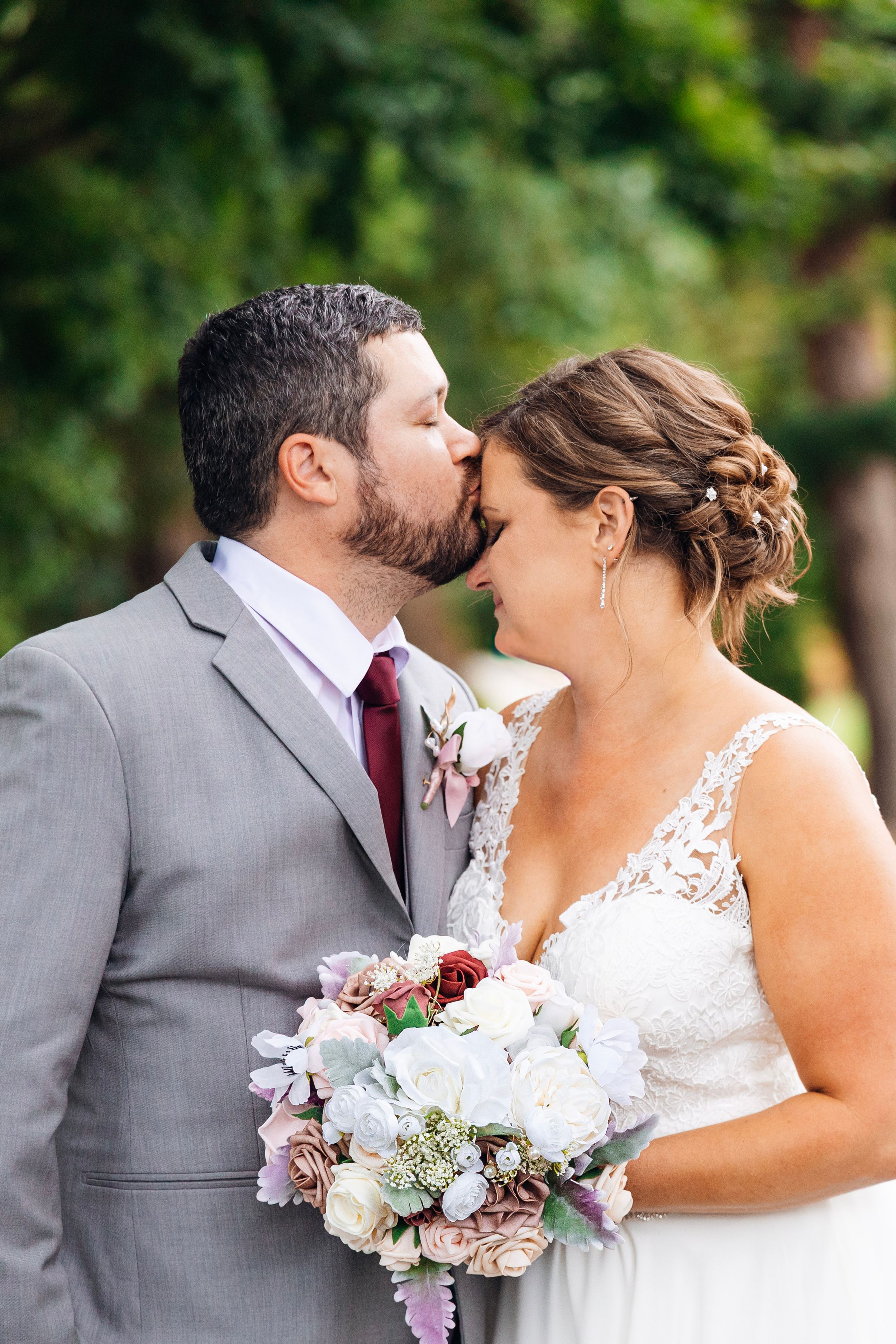 Bride and groom share a tender kiss outdoors, bride holding a bouquet of white, mauve, and burgundy flowers.