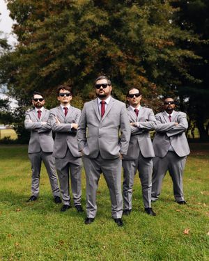 Five groomsmen in matching gray suits, red ties, and sunglasses pose confidently with arms crossed on a green lawn.