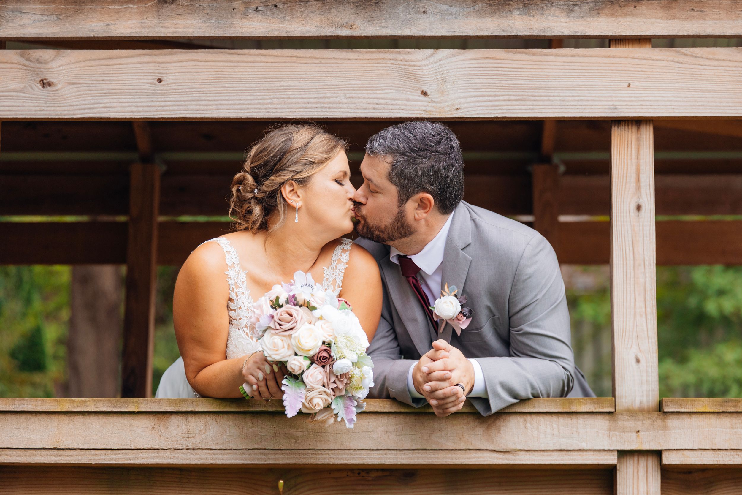 Bride and groom share a romantic kiss on a wooden bridge, bride holding a bouquet of white and pink flowers.
