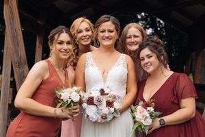 Bride in white lace gown poses with four bridesmaids in dusty rose and burgundy dresses holding floral bouquets.