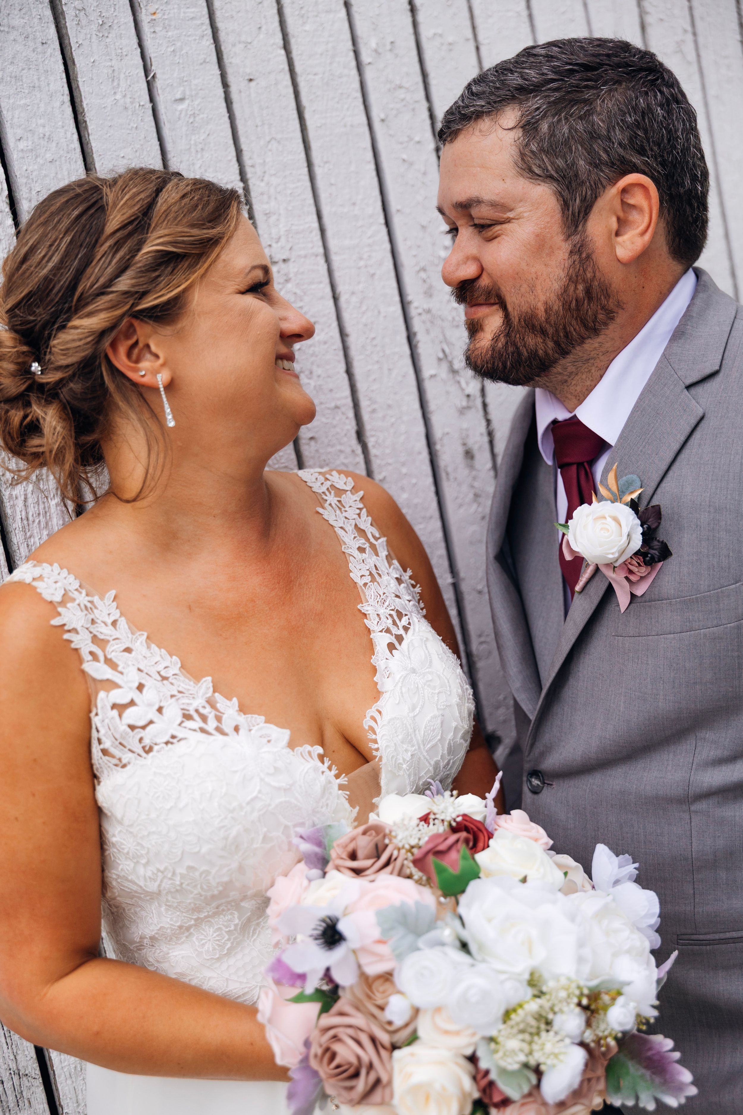 Bride and groom smiling at each other, bride holding a pastel bouquet, groom in gray suit with floral boutonniere.