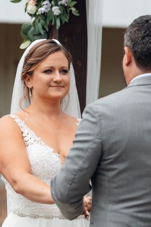 Bride in white lace gown and veil holds hands with groom in gray suit during outdoor wedding ceremony.