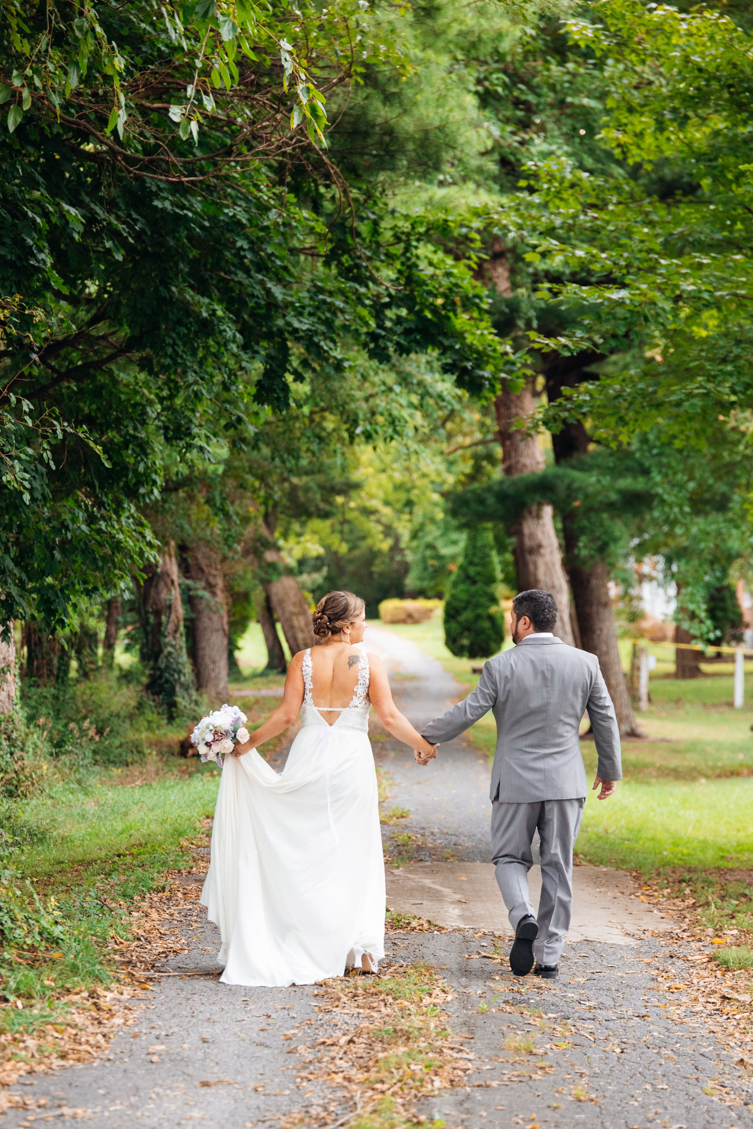 Bride and groom walking hand in hand down a tree-lined path, bride in white gown holding bouquet, groom in gray suit.