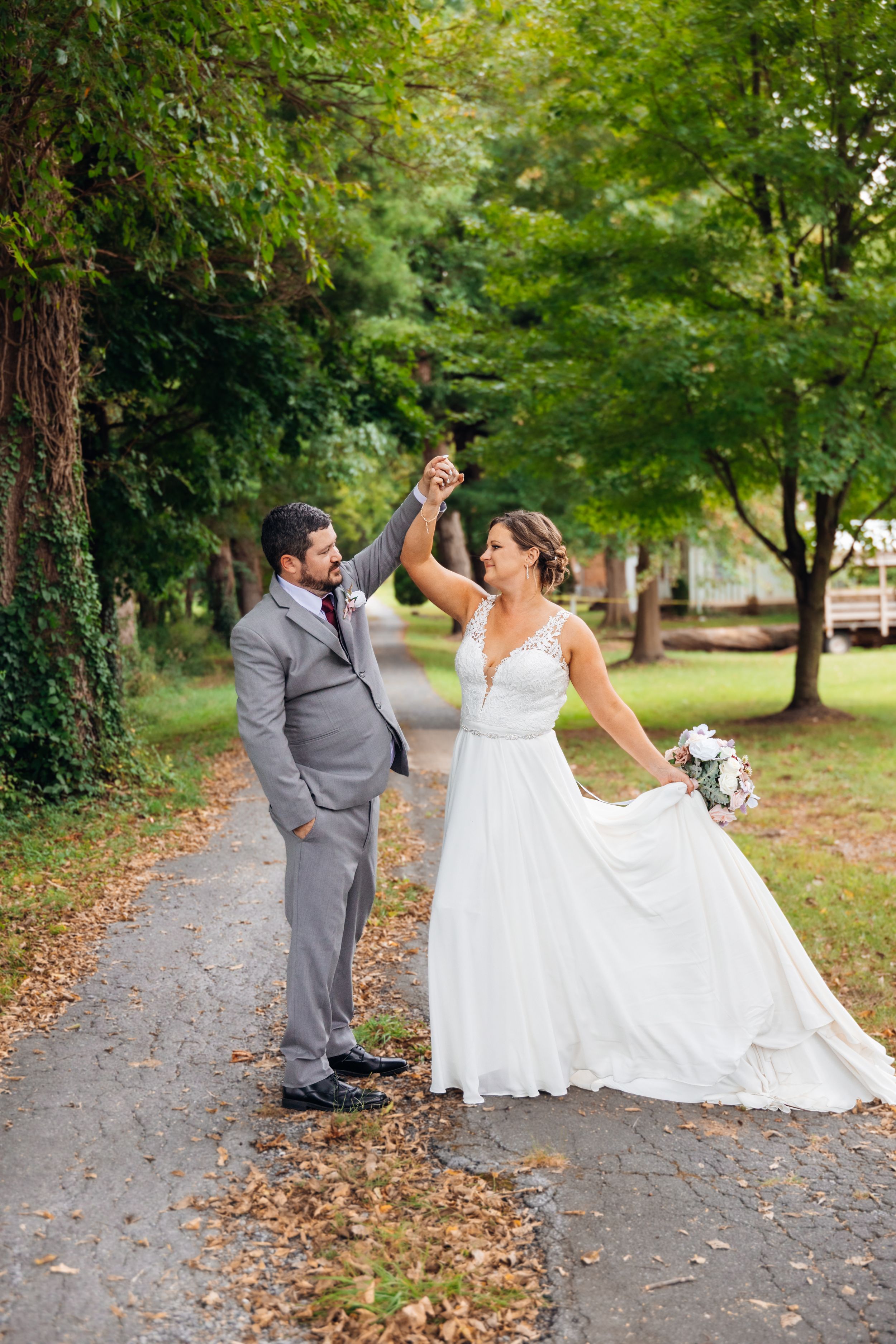Groom in gray suit twirling bride in white lace gown on a tree-lined path, holding bouquet of white flowers.