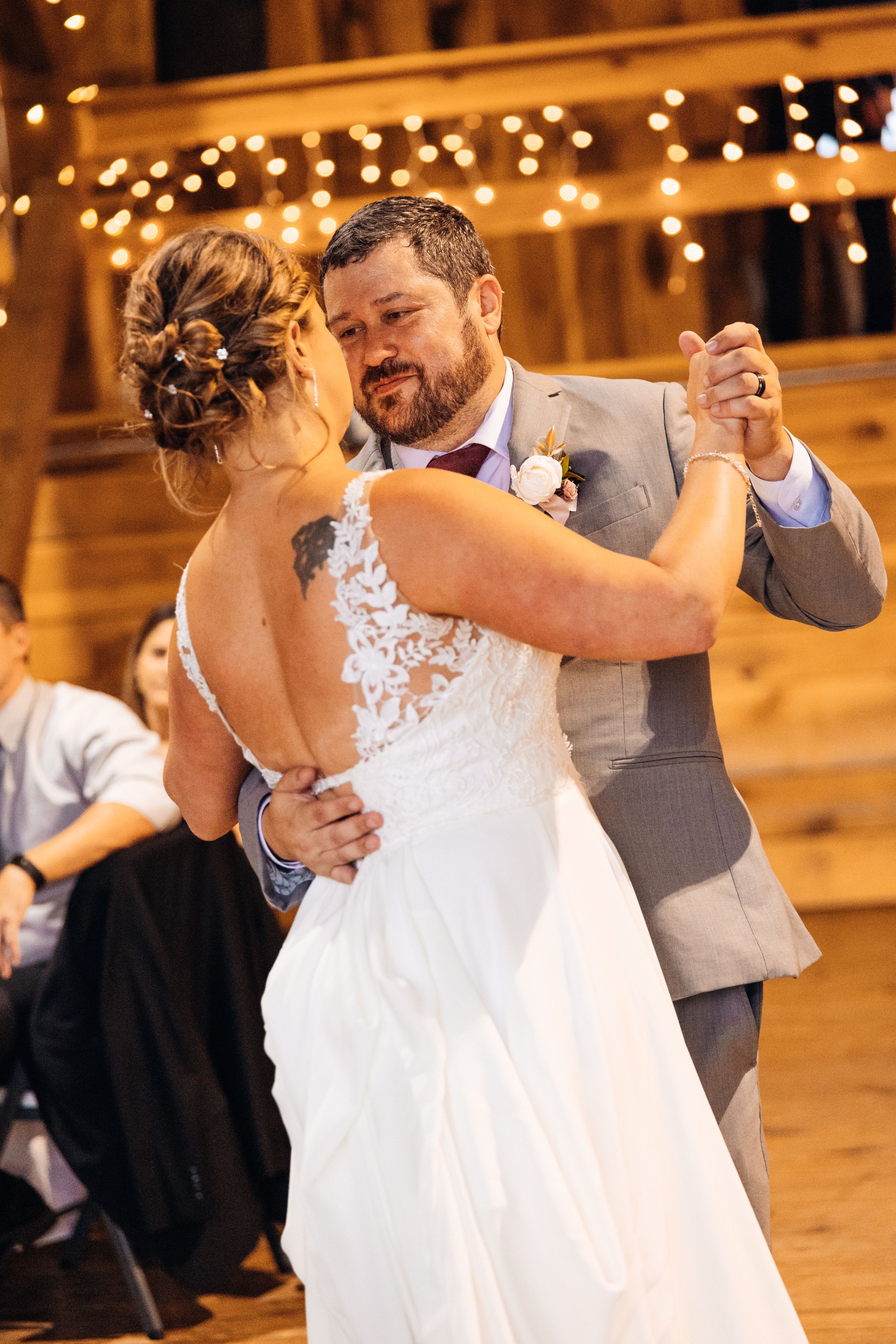 Bride and groom share their first dance in a rustic barn venue adorned with warm string lights during wedding reception.