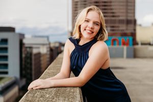 Smiling blonde woman in navy dress leaning on rooftop ledge with city skyline in background.