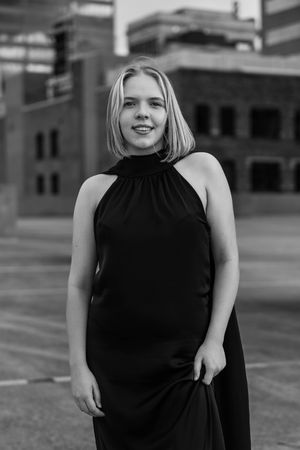 Young blonde woman in black halter dress poses confidently outdoors in urban setting, black and white portrait photo.