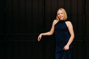 A smiling blonde woman in a navy halter dress poses elegantly against a dark wooden wall.