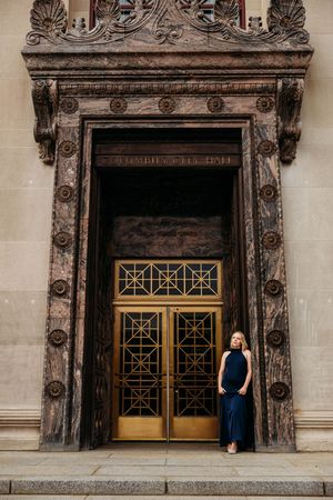 Woman in navy dress poses before ornate gold doors of a grand historic building with decorative stone facade.