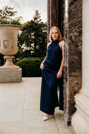 Woman in navy halter maxi dress posing by marble column on elegant terrace with stone urn and greenery.
