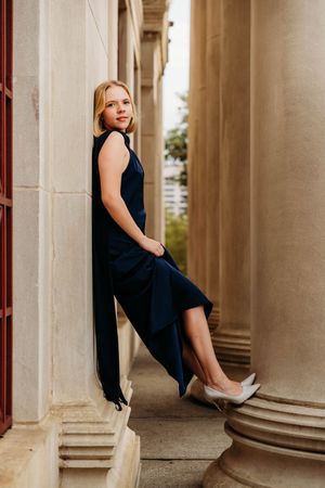 Woman in navy jumpsuit leaning against stone columns, wearing white heels, elegant outdoor portrait.