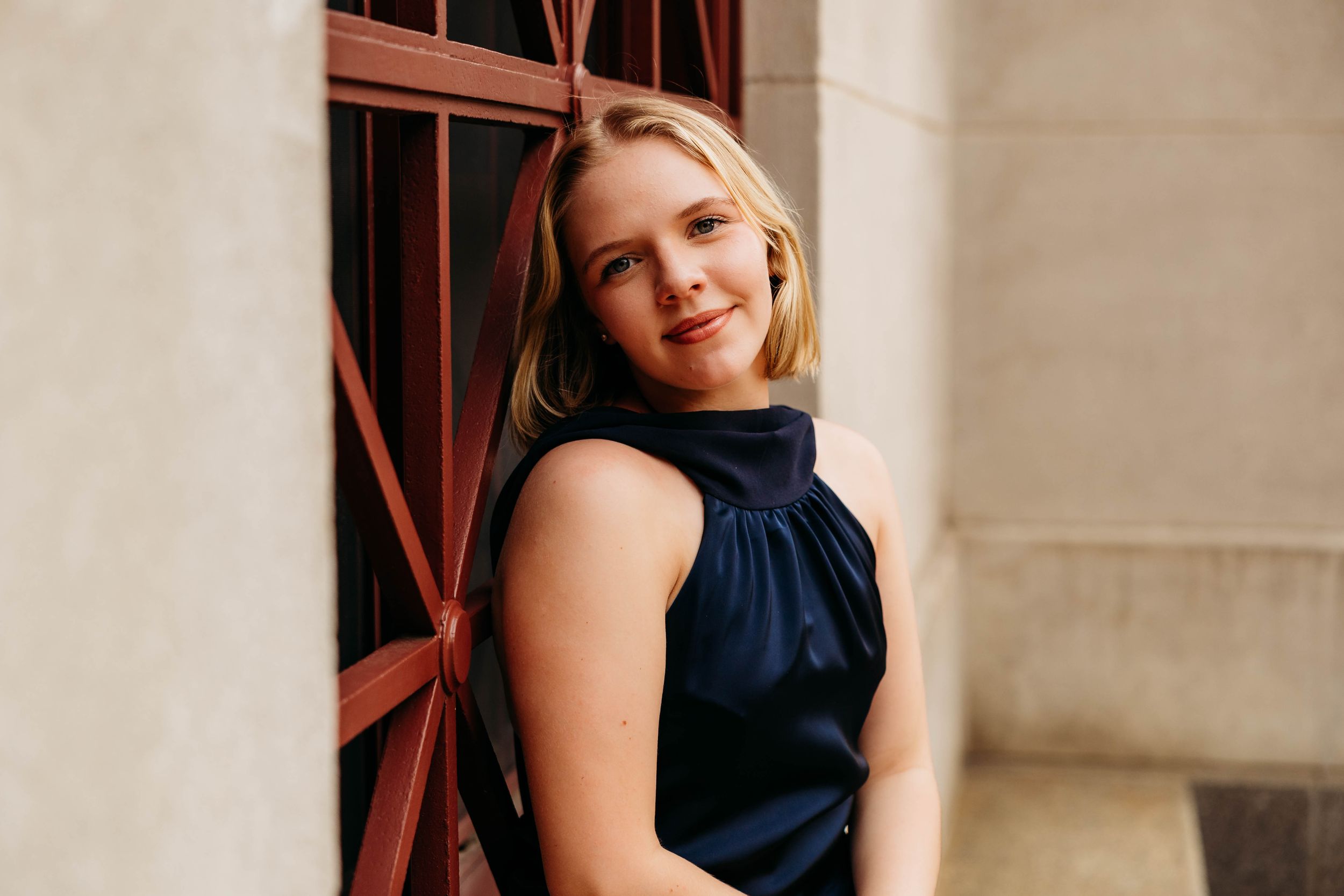 Young blonde woman in navy dress smiling over her shoulder, leaning against a red door outdoors.