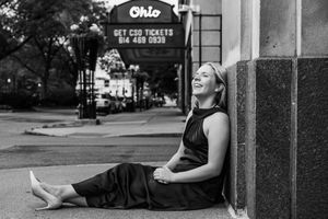 A woman in a black dress sits laughing against a building near the Ohio Theatre marquee in a black and white photo.