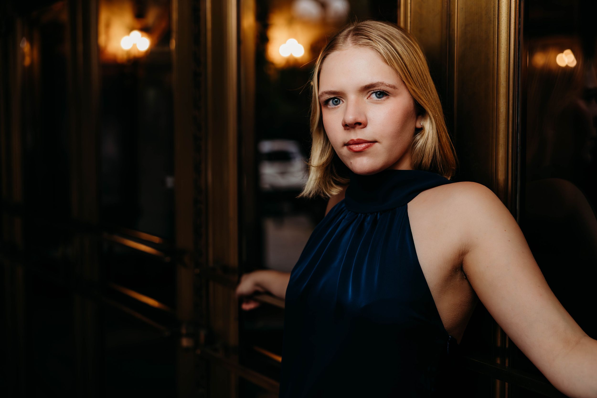 Elegant blonde woman in navy halter dress posing by golden doors in dramatic ambient lighting.