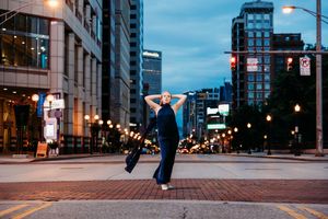 A woman in a navy jumpsuit poses gracefully on a city street at dusk with glowing lights and buildings.