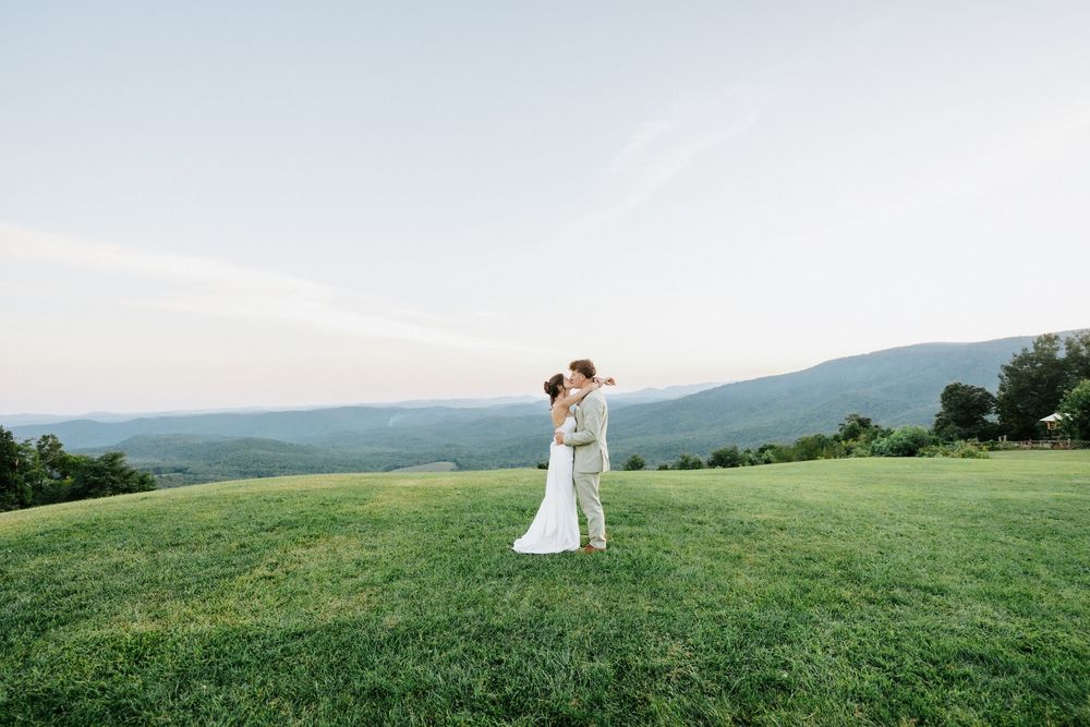 Bride and groom embrace on lush green hillside with sweeping mountain valley views under soft blue sky.