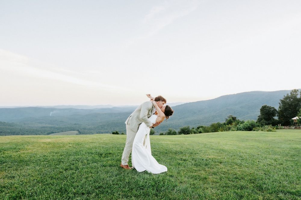 Groom dips bride on lush green hilltop with stunning mountain valley views at outdoor wedding ceremony.