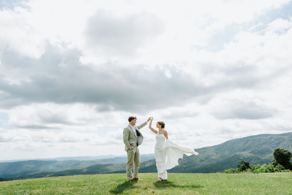 Bride and groom dancing on a scenic mountain hilltop under dramatic cloudy skies on their wedding day.