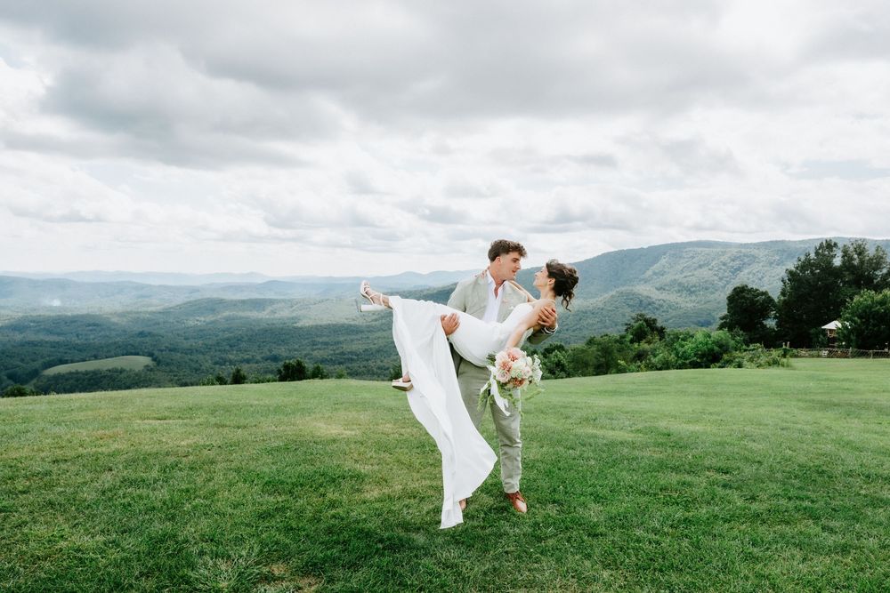 Groom lifts bride in white dress on green hilltop with scenic mountain valley backdrop on their wedding day.