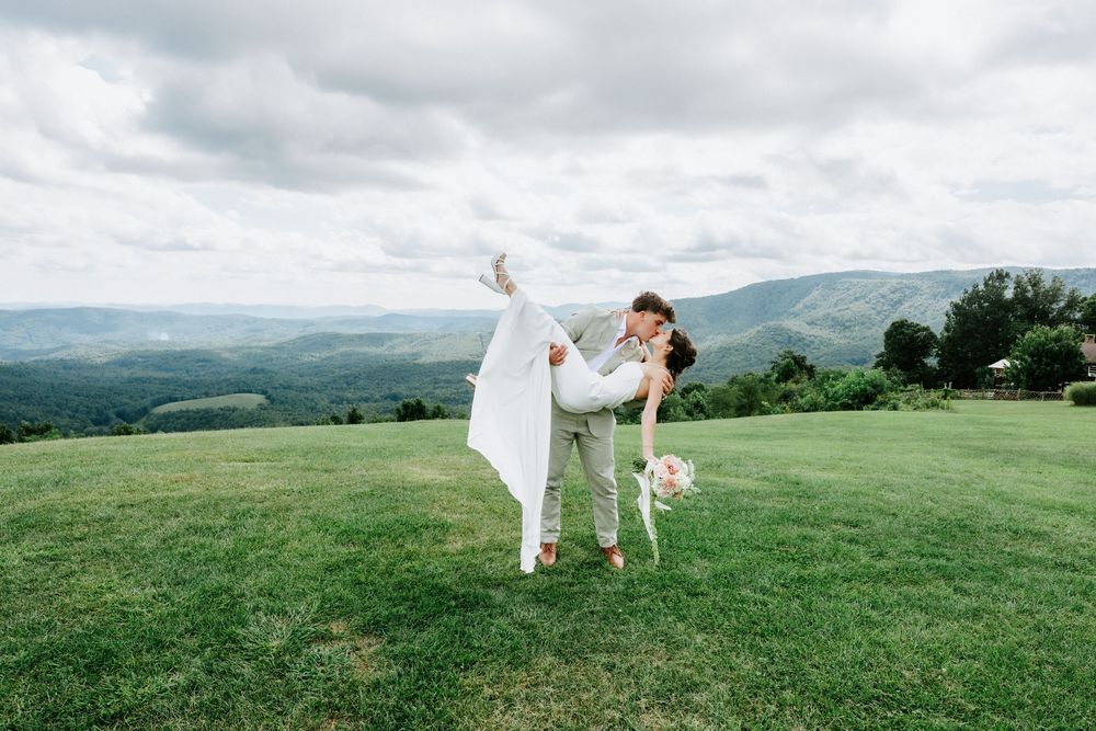 Groom lifts bride in white dress on green hillside with mountain views during romantic outdoor wedding photo.