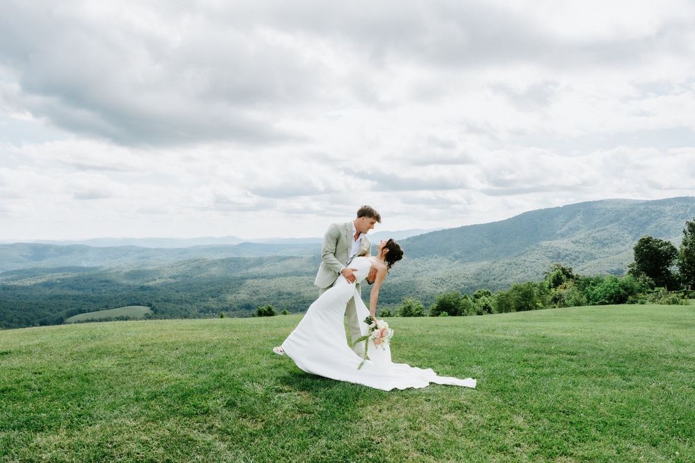 A groom dips his bride in a white gown on a lush green hilltop with stunning mountain views under a cloudy sky.