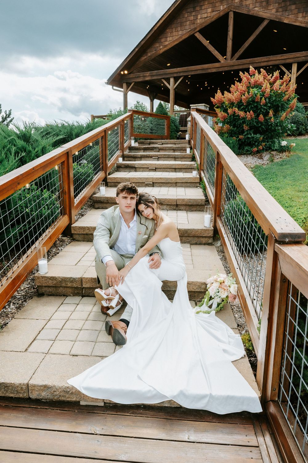 Bride and groom sitting on outdoor wooden staircase steps at wedding venue surrounded by lush greenery.