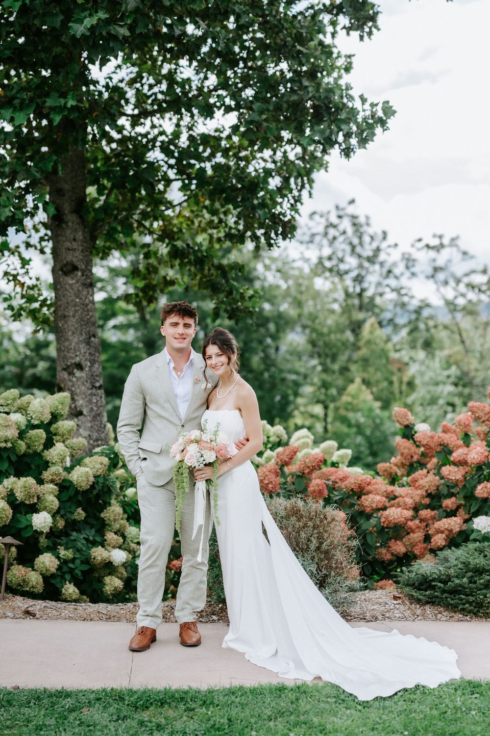 Wedding couple posing together in garden surrounded by blooming hydrangeas, bride holding bouquet.