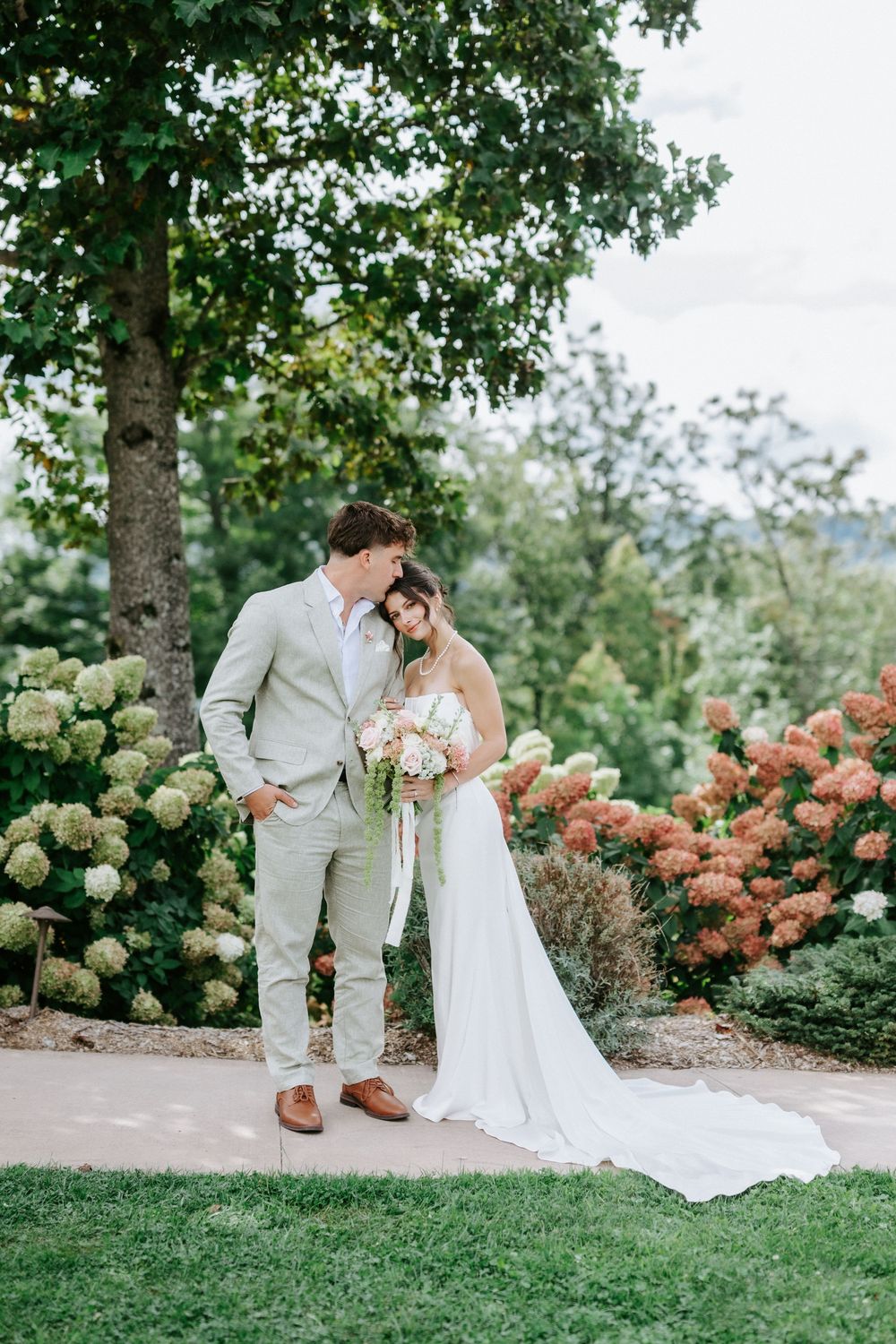 A bride and groom pose together in a lush garden with blooming hydrangeas, the bride holding a soft pink bouquet.