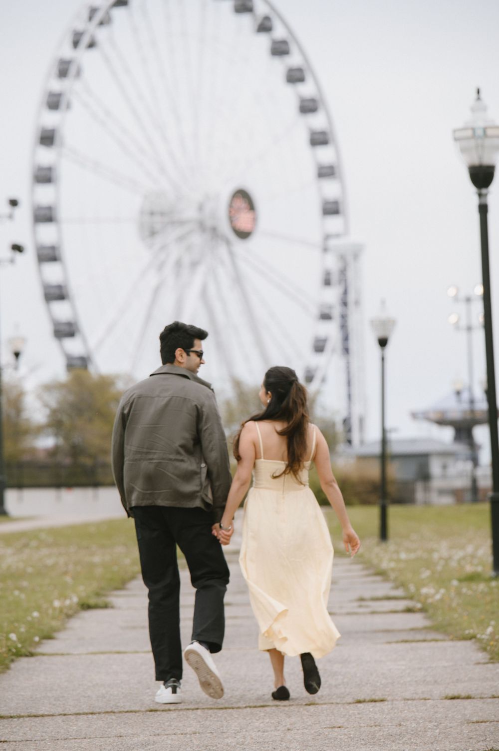 Couple holding hands walking toward a large Ferris wheel, woman in yellow dress and man in gray jacket.