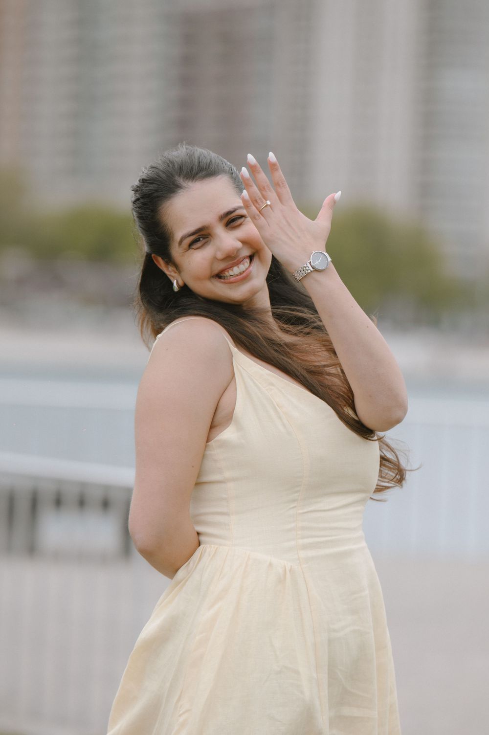 Smiling woman in a cream dress showing off a ring and watch while posing outdoors near a waterfront.
