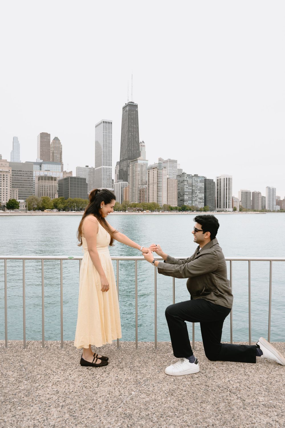 Man proposes to woman on one knee by Chicago lakefront with city skyline in background.